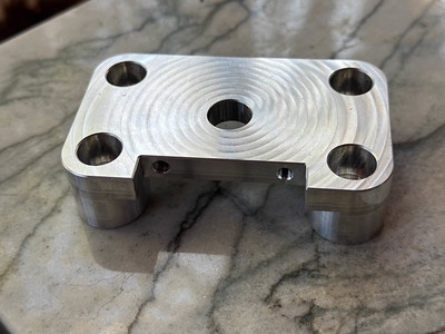 A shiny block of aluminum with four bolt holes and an extra hole in the middle. It has been machined to have a spiral pattern and the bolt holes in the front are visible in a recess