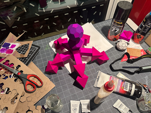 A colorful sculpture in reds, blues, and purples made up of geometric shapes in a form reminiscent of a bacteriophage. It has five-fold symmetry