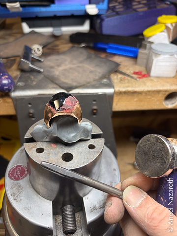 The ring is partly covered by red wax. It's being held in a vise attached to the bench I'm working at. Gray melted-looking plastic holds the bottom of the band to be gripped in the vise.