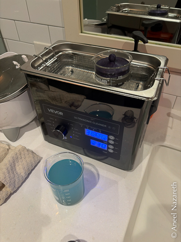 An ultrasonic cleaner sitting on the counter in my bathroom. It is a blocky machine with a basin of water in the top. In the water is a beaker with a lid on it. On the counter is another beaker filled with blue liquid. The blue is from the copper in the bronze reacting with the vinegar.